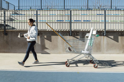 Side view of young female athlete carrying starting blocks while walking near cart with gear before track and field training on sunny day on stadium