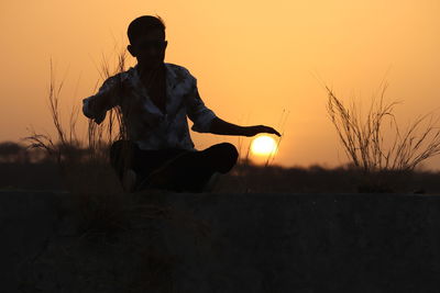 Optical illusion of man protecting sun while sitting on field against sky during sunset