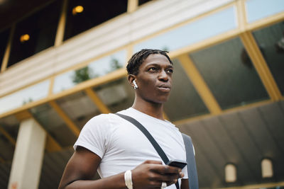 Young male student listening music through wireless in-ear headphones in university campus