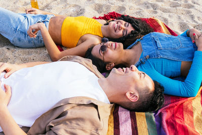 Diverse man and women in casual clothes lying on blanket and smiling while spending time on sandy beach near sea on summer weekend day