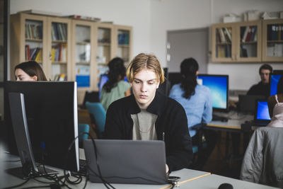 Young male high school student using laptop at desk against teacher and friends sitting in computer lab