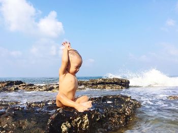 Full length of boy sitting on rock at sea against sky