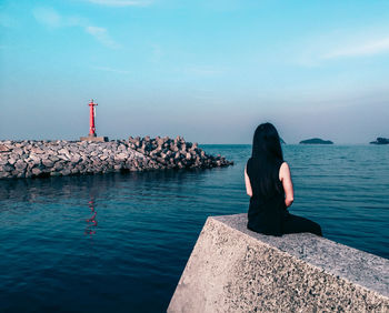 Rear view of woman sitting on retaining wall by sea against sky