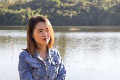 Thoughtful young woman looking away while standing against lake in forest