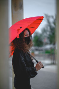 Woman with a mask and a red umbrella seen between two columns