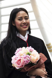 Portrait of smiling woman in graduation gown holding bouquet