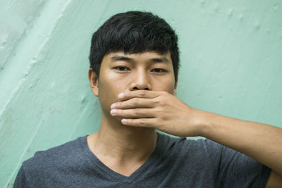 Close-up portrait of young man with hand covering mouth standing against wall