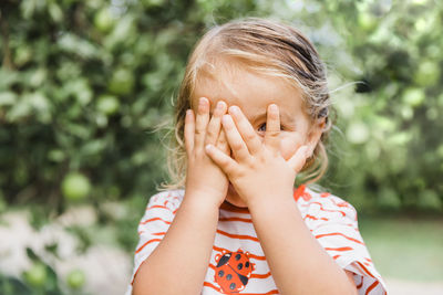 Close-up of cute girl hiding face with hands while standing against plants in park