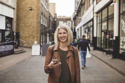 Portrait of smiling young woman holding mobile phone on road in city