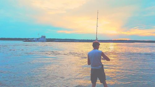 Rear view of man fishing on beach against sky during sunset