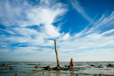 Full length of woman standing by dead tree on shore at beach against sky