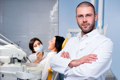 Portrait of confident male doctor with coworker operating patient in background at dental clinic