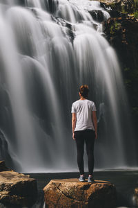 Rear view of man standing on rock looking at waterfall