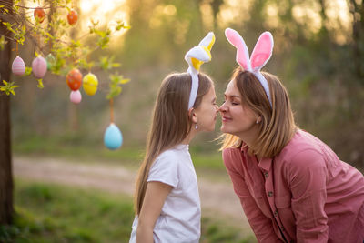 Mother and daughter standing by hanging easter eggs on branch at park