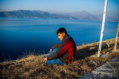 Thoughtful man sitting at lakeshore against mountains