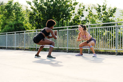 Full body of determined young multiethnic sportswomen doing squat exercise with resistance band during fitness workout in park in summer day