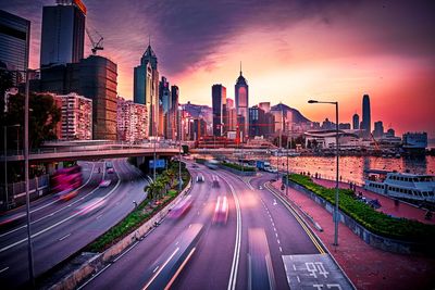 Light trails on road amidst buildings against sky during sunset