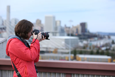 Rear view of woman photographing in city against sky during winter
