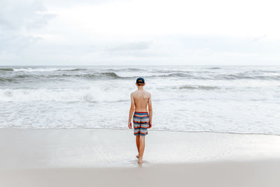 Rear view of teenage boy standing on beach against sky