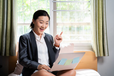 Young woman using phone while sitting on window