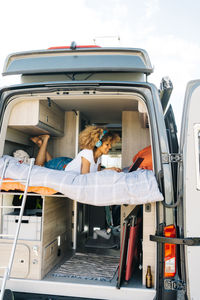 Cheerful african american female smiling and listening to music in headphones while lying on bed in caravan and browsing social media on laptop