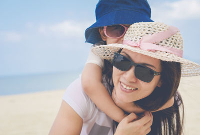 Close-up of smiling mother carrying daughter while standing at beach against sky