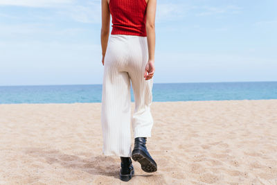 Back view of anonymous female in boots walking on sandy shore towards calm blue sea on sunny day