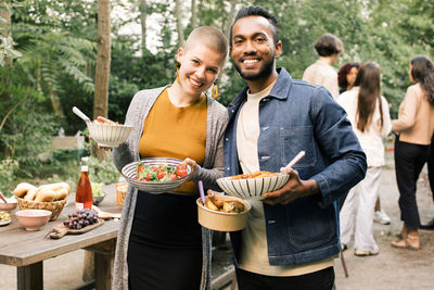 Portrait of smiling friends holding food bowls during dinner party in back yard