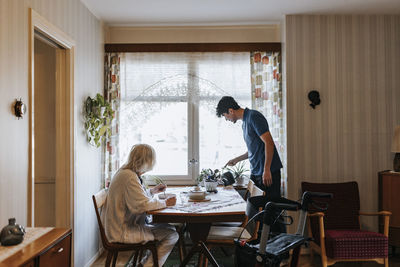 Male healthcare worker watering plants while senior woman eating food at home