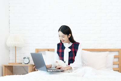 Young woman using phone while sitting on bed against wall