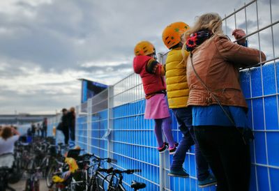 Spectators climbing on fence against cloudy sky