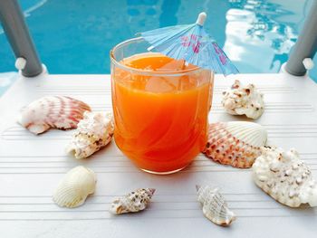 Glass of orange juice with blue umbrella and seashells near the blue pool water