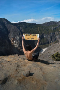 Rear view of shirtless man holding information sign while sitting on mountain