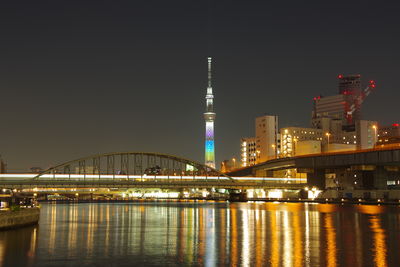 Illuminated bridge over river by buildings against sky at night