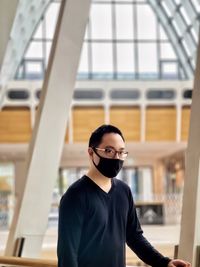 Young asian man in eyeglasses and face mask standing against skylight and architectural features.