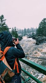 Rear view of person photographing while standing by railing on bridge over river