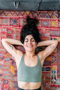 Top view of cheerful hispanic woman in sports bra holding hand behind head and smiling with closed eyes while lying on ornamental carpet near sofa during break in training at home