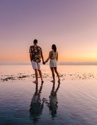 Rear view of couple standing at beach against sky during sunset
