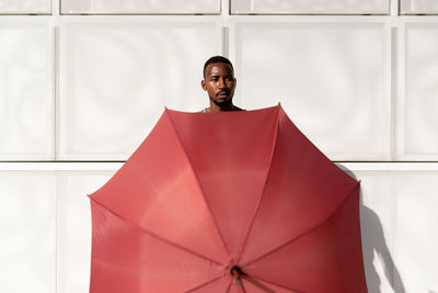 Black male standing on sidewalk in city and covering body with umbrella while looking at camera