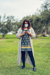 Full length of woman wearing mask holding phone standing at park