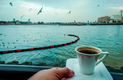 Cropped hand holding coffee cup against lake in city