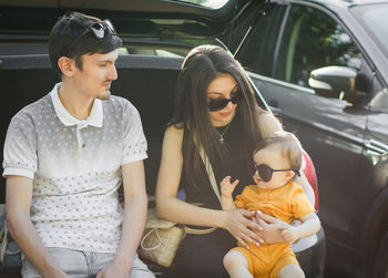 Caucasian young parents hold in their arms a small daughter in her arms sitting in the open trunk