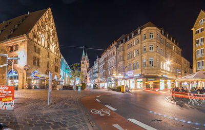 Illuminated city street by buildings against sky at night