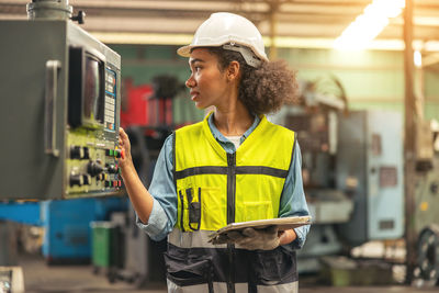 Standing in front of a control panel, a female industrial electrical engineer with a safety hardhat.