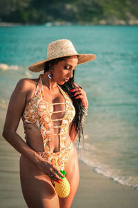 Seductive young woman wearing swimwear and hat while standing on shore at beach