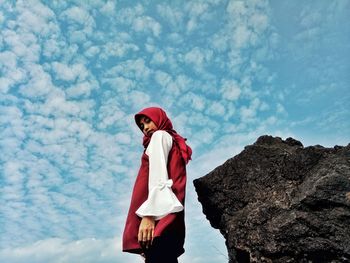 Low angle portrait of girl wearing hijab standing by rock against cloudy sky