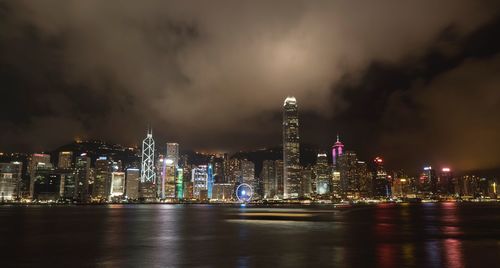 Illuminated city at waterfront against cloudy sky