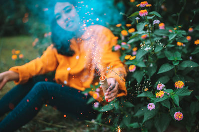 Woman holding lit sparkler while sitting by flowering plants