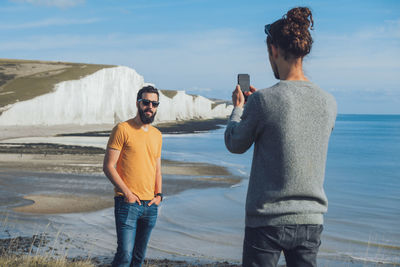 Young man photographing while standing on sea shore against sky