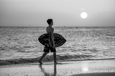 Side view of shirtless teenage boy holding surfboard walking at beach against sky during sunset